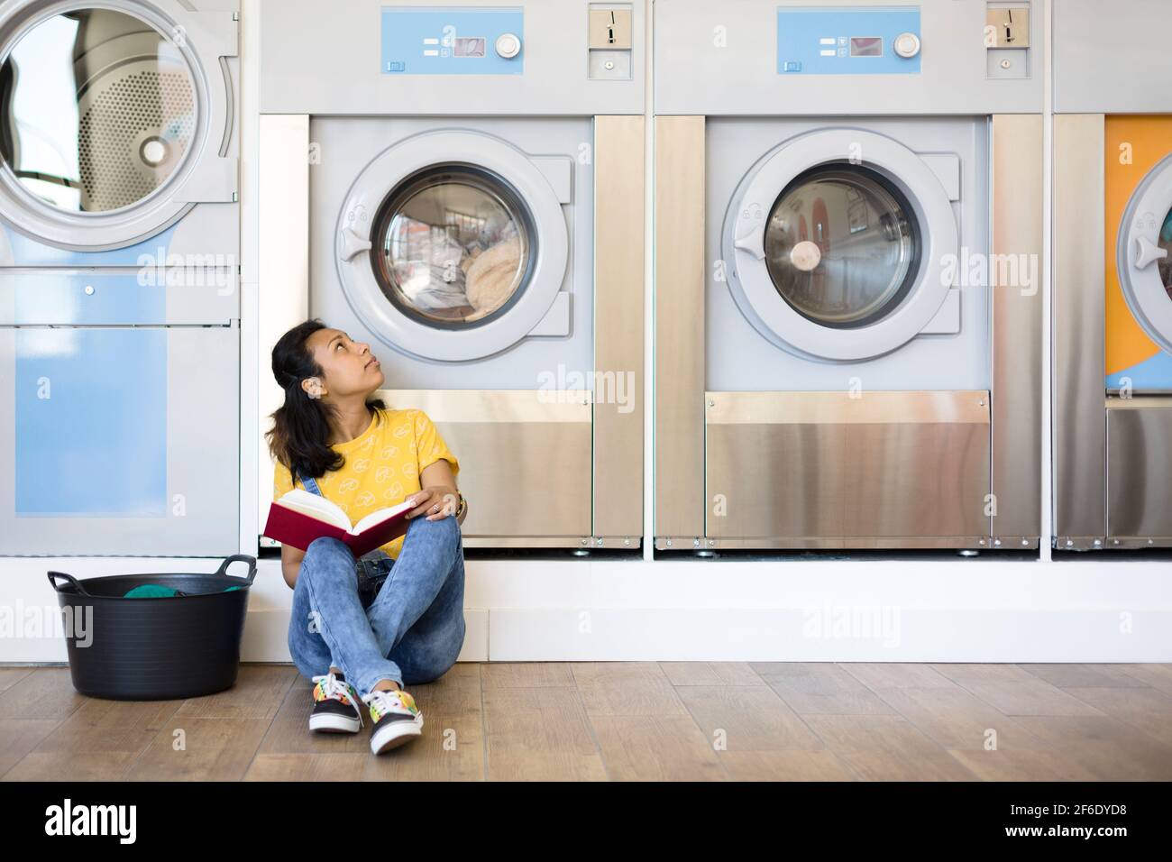 Donna ispanica seduta sul pavimento in attesa di lavare i vestiti nella lavanderia self-service. Spazio per il testo. Foto Stock
