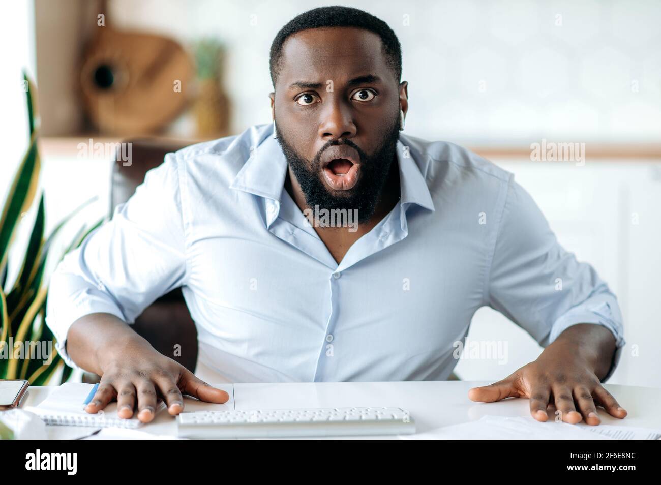 Un ragazzo alfiere afro-americano sorpreso che indossa una camicia formale, ha ricevuto notizie inaspettate, stupito guardando la macchina fotografica mentre si siede sul posto di lavoro Foto Stock