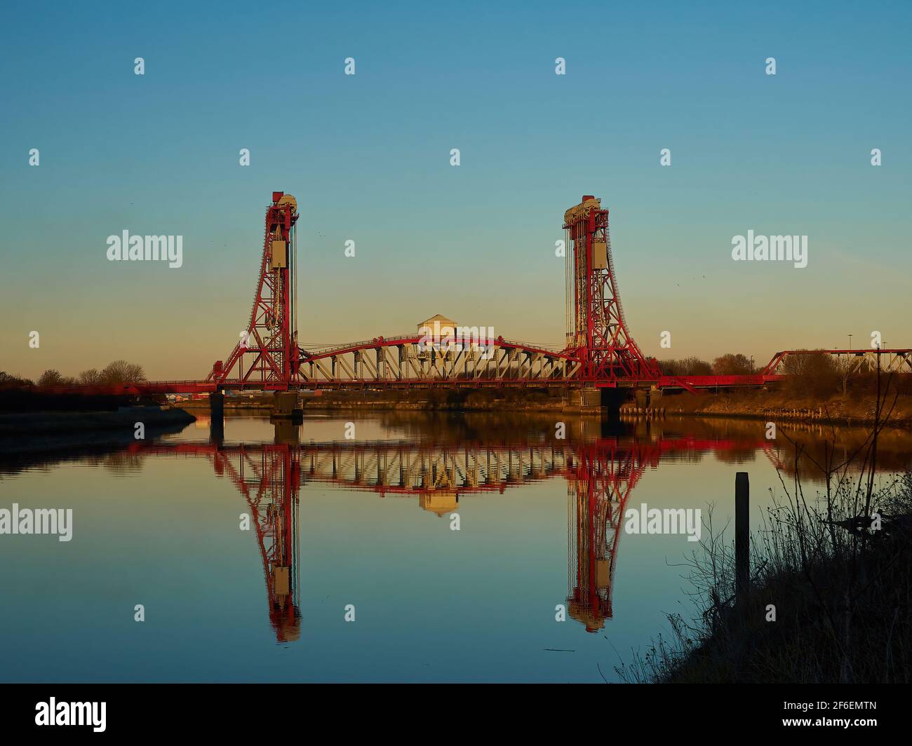 Il Newport Bridge contro un cielo blu e arancione classificato. Il primo piano mostra le Tee del fiume ancora e vetrose che riflettono la scena e il cielo. Foto Stock