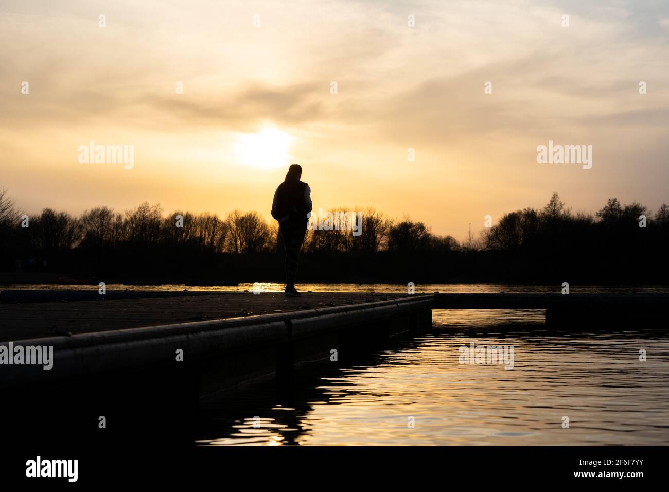 Donna in piedi da sola al tramonto alla fine del molo del lago pensieroso e tranquillo fiume sotto. Aurea alba estate peccato luce che si riflette sull'acqua solo Foto Stock