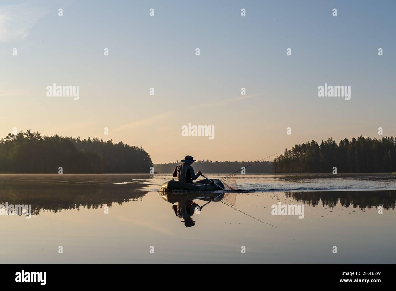Pescatore in una barca gonfiabile che si candida su un tranquillo lago forestale, all'alba, sullo sfondo di cielo e alberi. Bellissimo paesaggio. Foto Stock