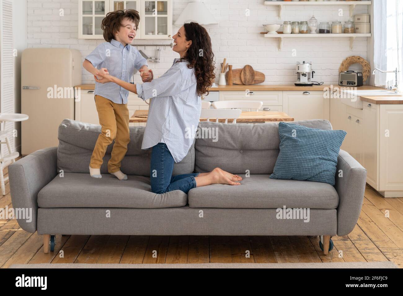 Madre e bambino che ride divertendosi, tenendo le mani e saltando sul divano a casa. Buona famiglia. Foto Stock