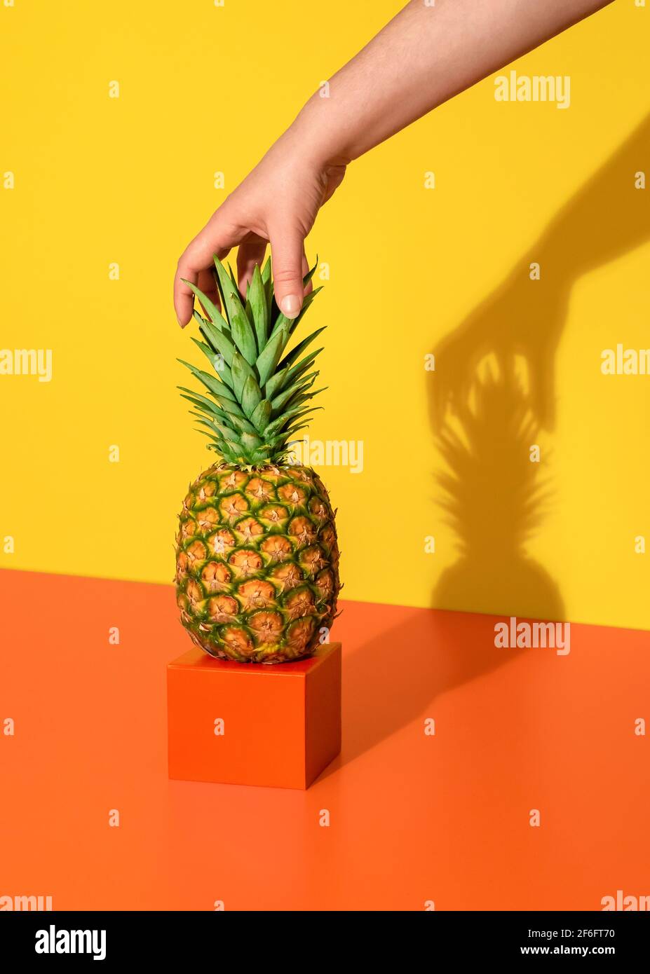 La mano della donna afferra l'ananas dalle foglie. Immagine minima con ananas singolo su un tavolo arancione su sfondo giallo. Foto Stock