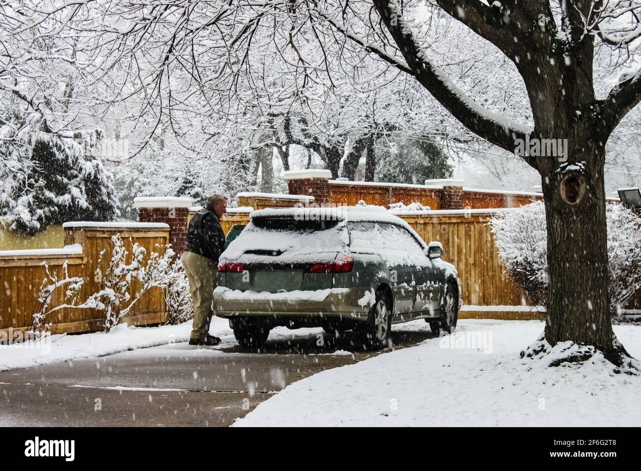 Uomo di mezza età con auto coperta di neve in vialetto di accesso giornata estremamente nevosa con neve che cade Foto Stock
