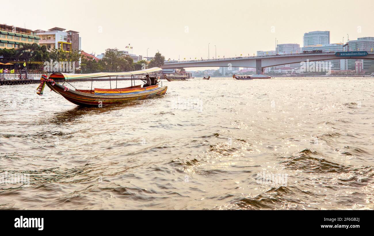 Barca tradizionale a coda lunga Thailandia Barca a coda lunga con turisti che passano lungo il fiume Chao Phraya nel pomeriggio, circondato dallo sfondo Foto Stock