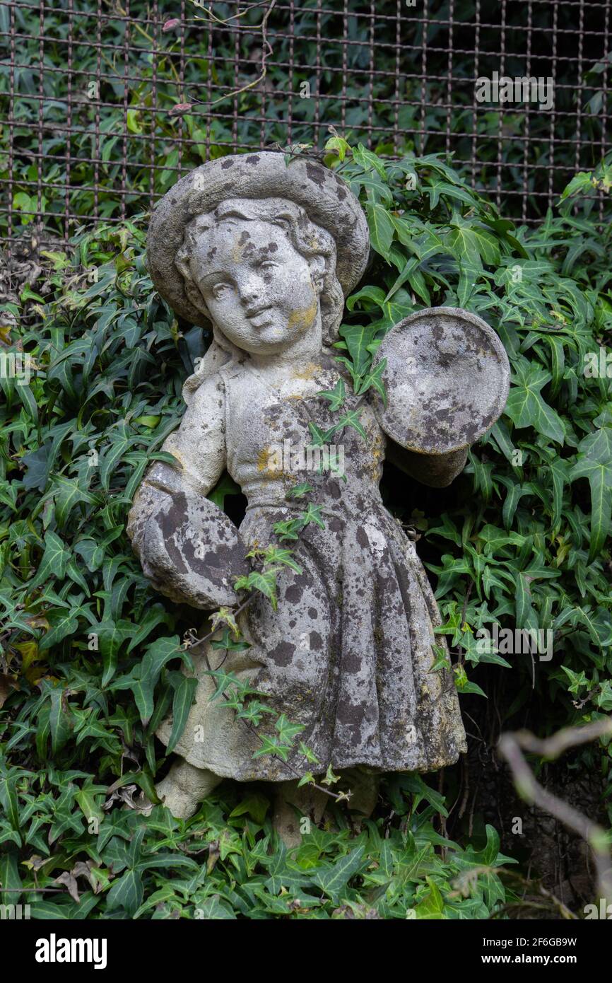 Vecchia statua bianca intemperie di una ragazza che gioca Clash Cymbals circondato da edera Foto Stock