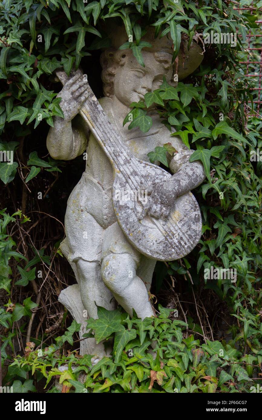 Vecchia statua bianca intemperie di un ragazzo che suona un liuto circondato da edera Foto Stock