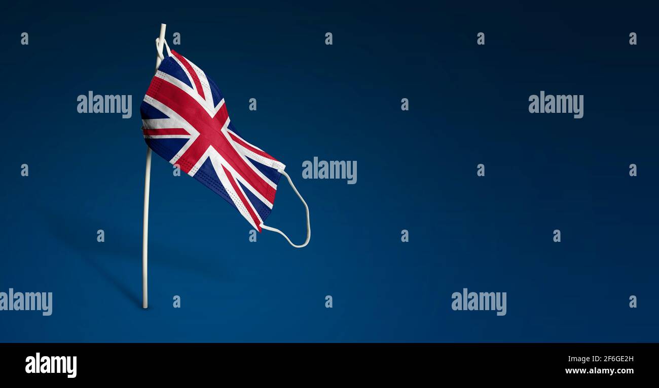 Maschera per il Regno Unito su sfondo blu scuro. Bandiera ondulata del Regno Unito dipinta su maschera medica sul palo. Flag di attacco del virus. Concetto della bandiera della lotta Foto Stock
