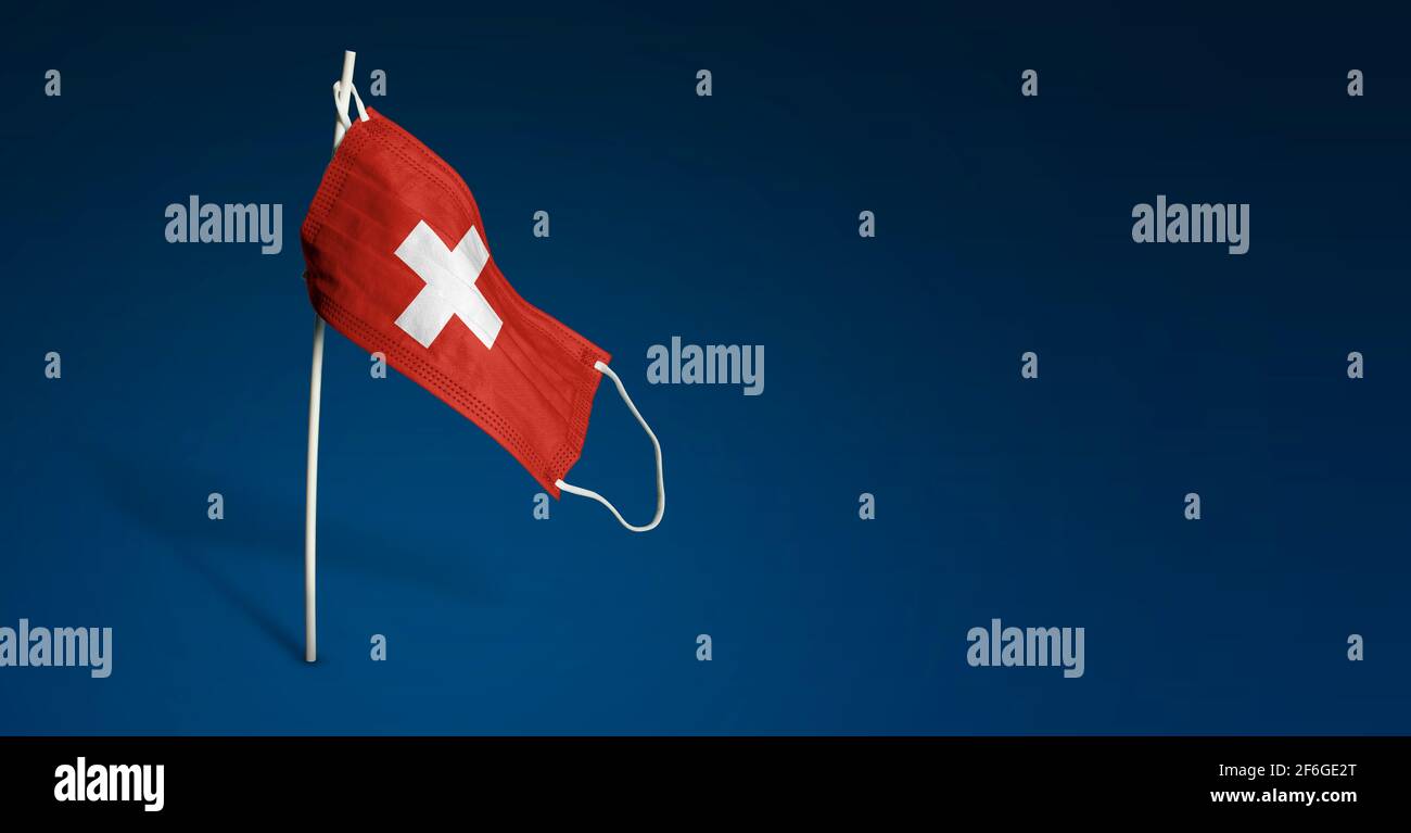 Maschera Svizzera su sfondo blu scuro. Bandiera sventolante della Svizzera dipinta su maschera medica sul palo. Flag di attacco del virus. Concetto del banner del Foto Stock