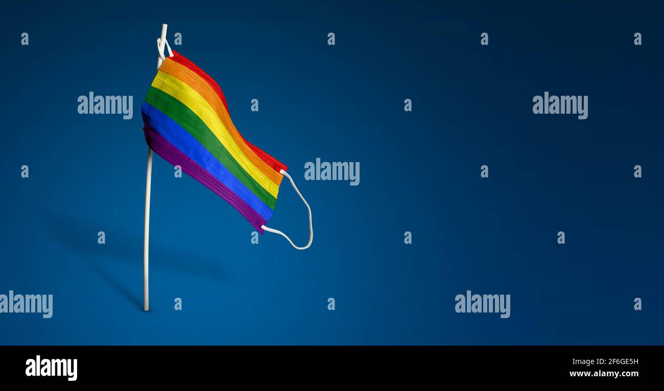 Maschera LGBT su sfondo blu scuro. Bandiera ondulata di LGBTQ dipinta su maschera medica sul palo. Flag di attacco del virus. Concetto della bandiera della lotta contro Foto Stock