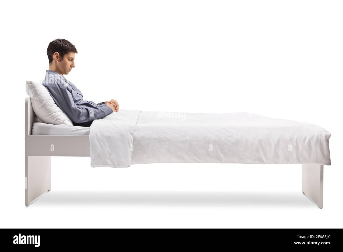 Uomo assonnato in pigiama seduto su un letto singolo sotto piumone isolato su sfondo bianco Foto Stock