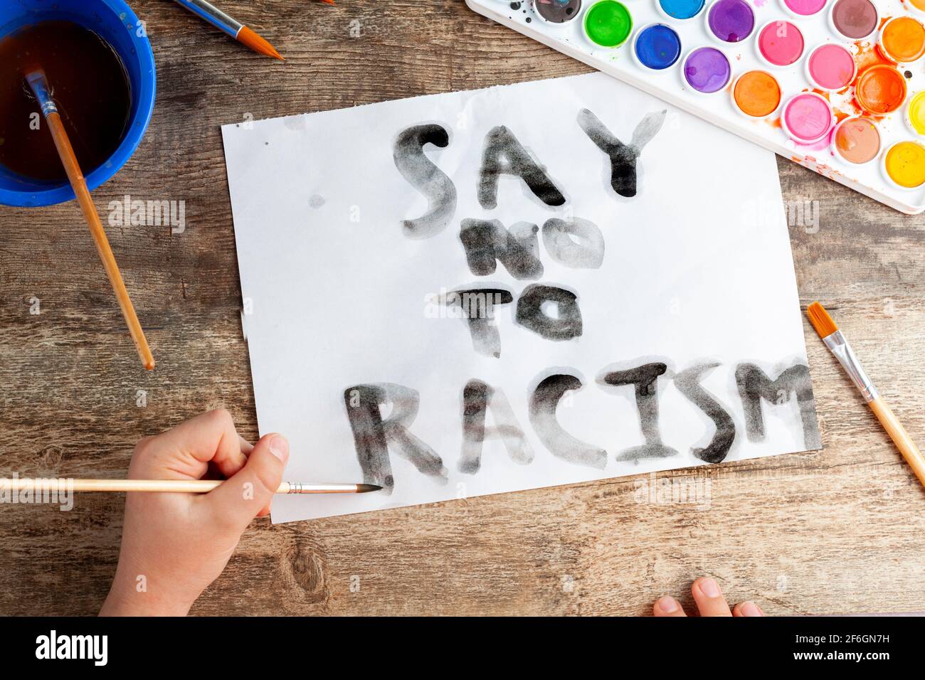 Il bambino caucasico sta scrivendo uno slogan di 'dire no al razzismo' usando il pennello e l'acquerello nero su un foglio bianco. Un bambino che è cosciente delle questioni sociali Foto Stock