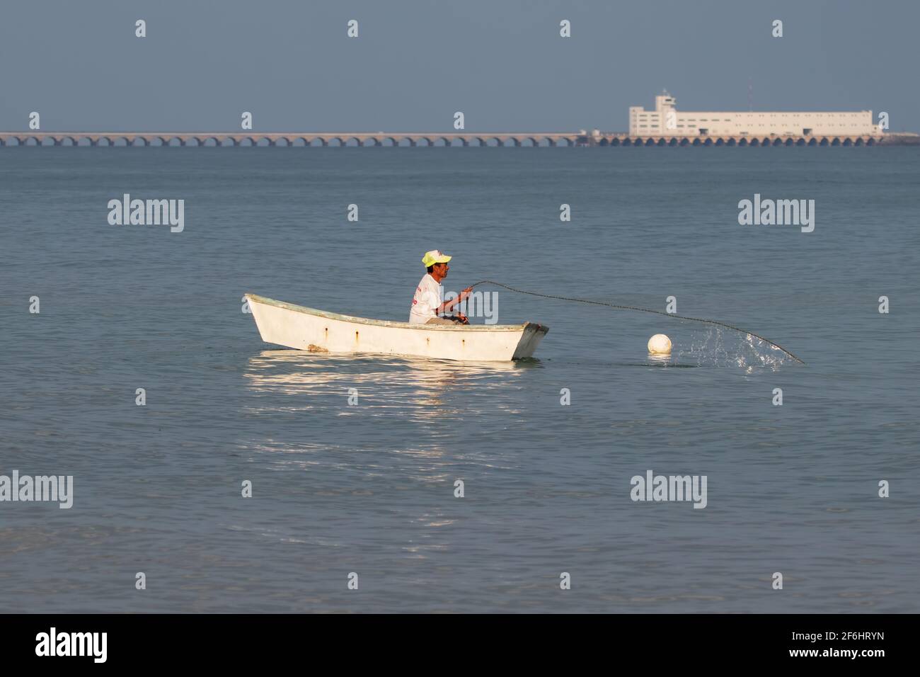 Pescatore in una piccola barca che tira nelle sue reti, un lungo molo commerciale è sullo sfondo - mare blu calmo in una giornata limpida Foto Stock