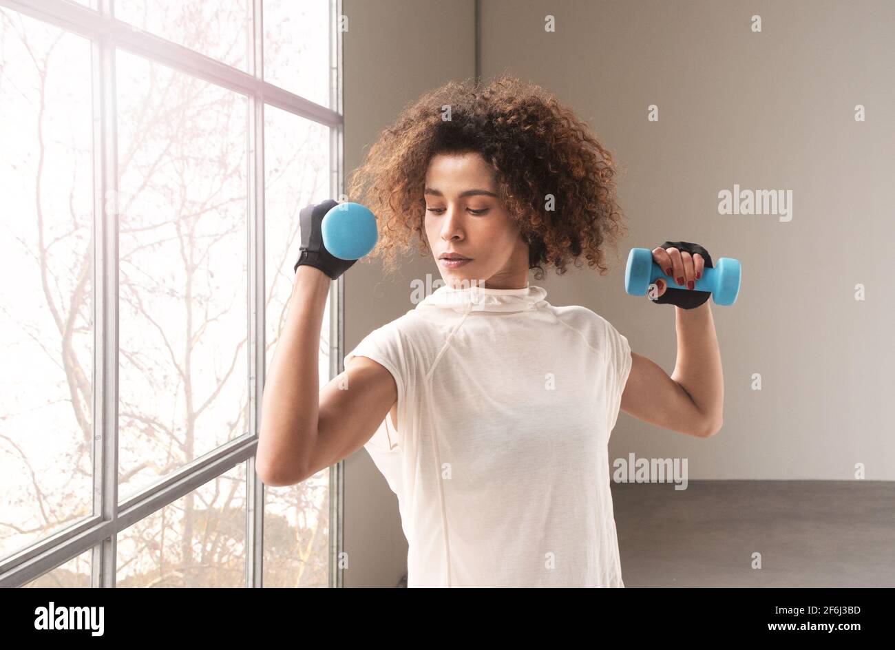 primo piano della cima di un giovane e bello donna che fa l'allenamento con manubri vicino alla finestra che indossa una maglietta bianca orizzontale alberi sfondo Foto Stock