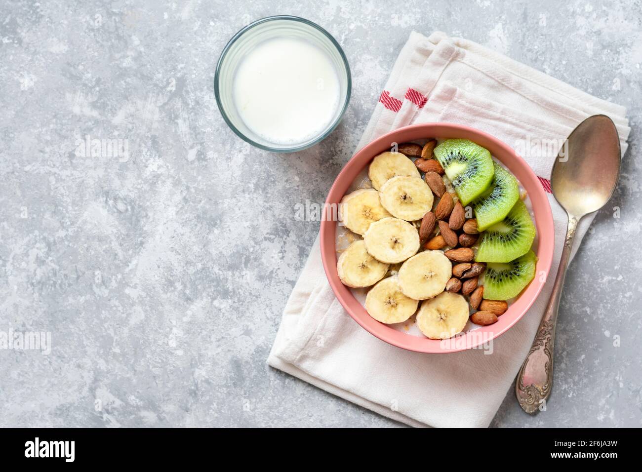 Farinata d'avena con banana, mandorle, noci e fette di kiwi in ciotola rosa, bicchiere di latte, tovagliolo su fondo grigio in cemento Vista dall'alto colazione o pranzo sani Foto Stock