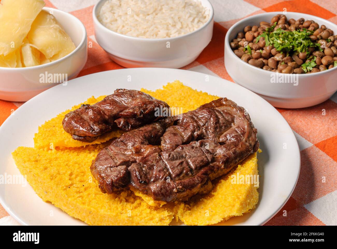 Carne arrosto con couscous brasiliano, abbinato a riso, fagioli, manioca e vinaigrette, su un piatto bianco sopra un tovaglia a scacchi bianco e arancione Foto Stock