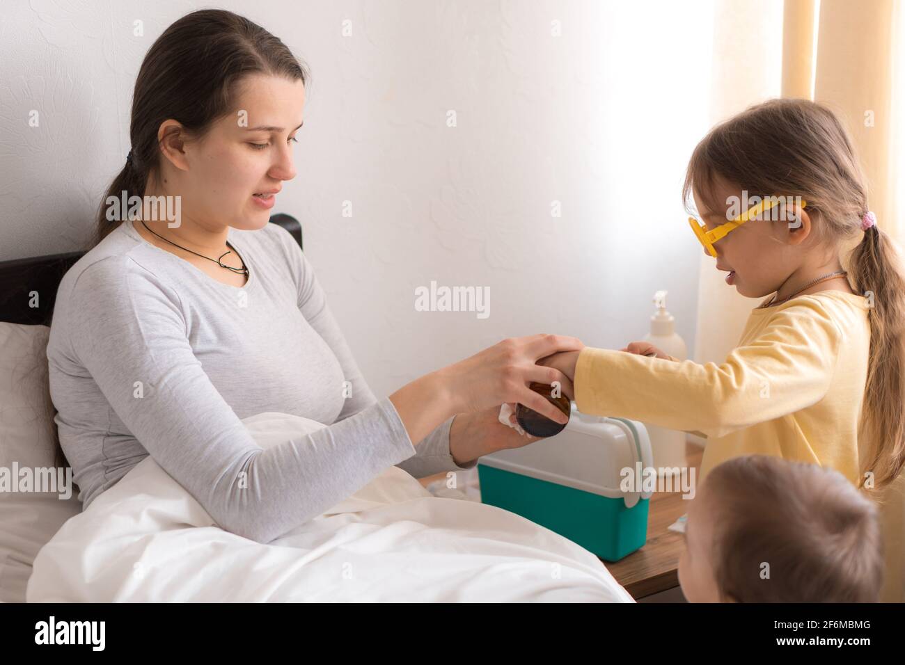 Carino bambina che indossa uniforme medico o infermiera con mamma malata o panna in camera da letto, dà a bere la medicina sciroppo di tosse, prendersi cura della famiglia Foto Stock