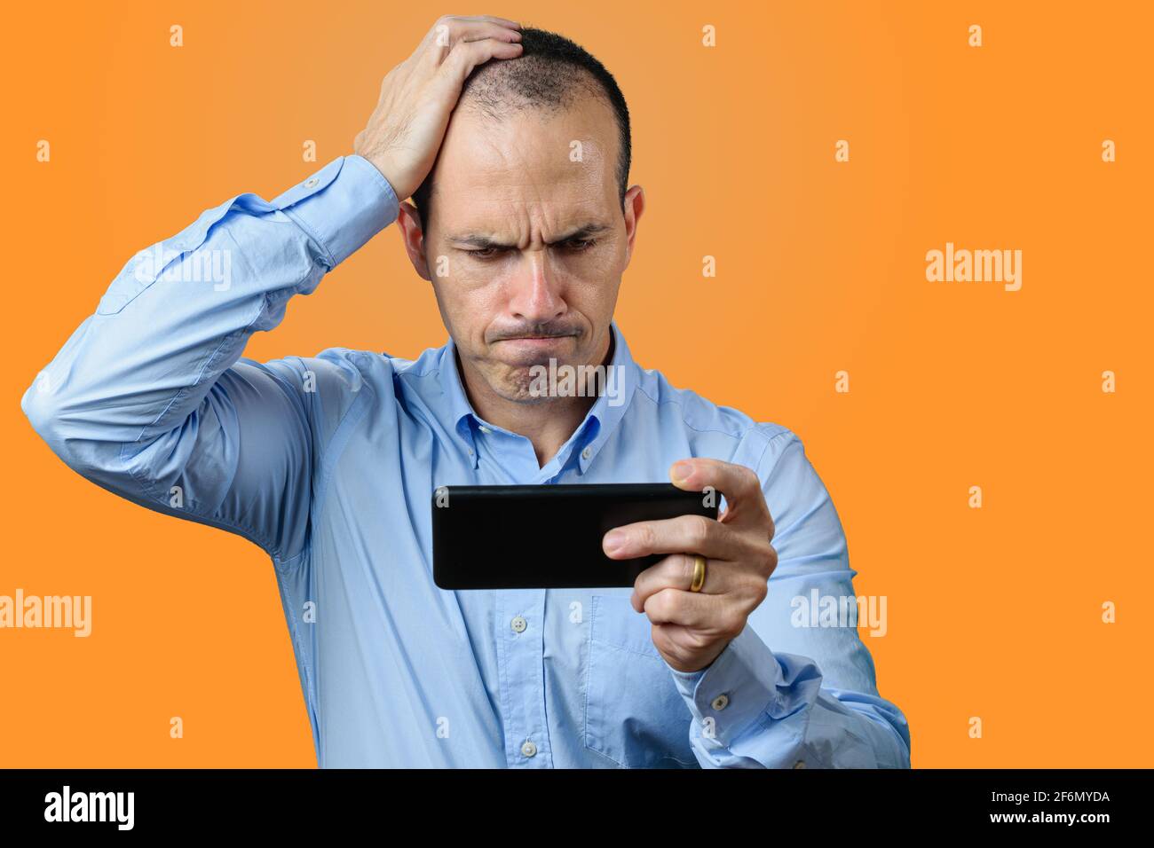 Uomo maturo in abbigliamento formale, guardando il suo smartphone, deluso e con la mano destra sulla testa. Sfondo arancione. Foto Stock