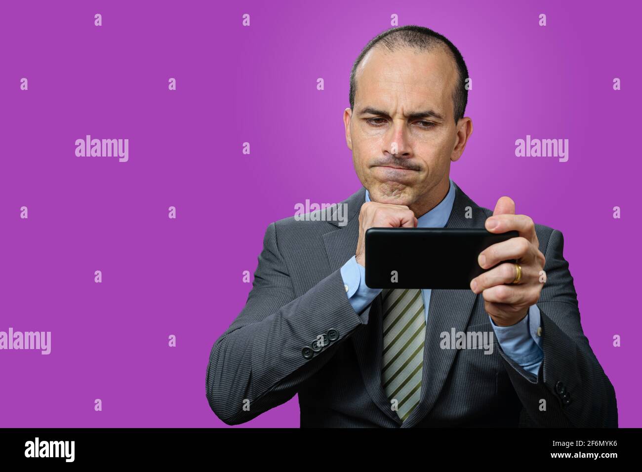 Uomo maturo con vestito e cravatta, guardando il suo smartphone, deluso e con il pugno sotto il mento. Sfondo viola. Foto Stock
