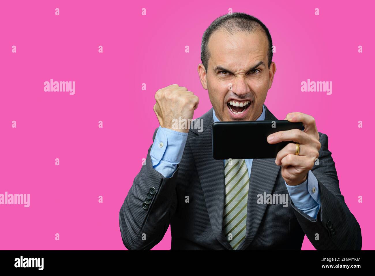 Uomo maturo con abito e cravatta, tenendo il suo smartphone orizzontalmente e festeggiando con un pugno chiuso. Sfondo rosa. Foto Stock