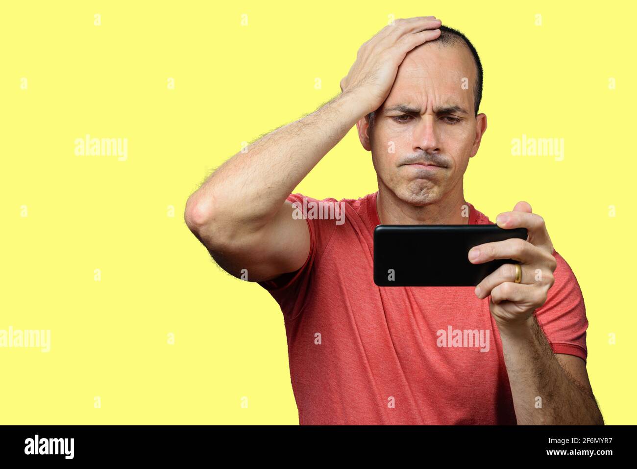 Uomo maturo in abiti casual, deluso, con la mano distesa sulla testa e tenendo lo smartphone orizzontalmente. Sfondo giallo. Foto Stock