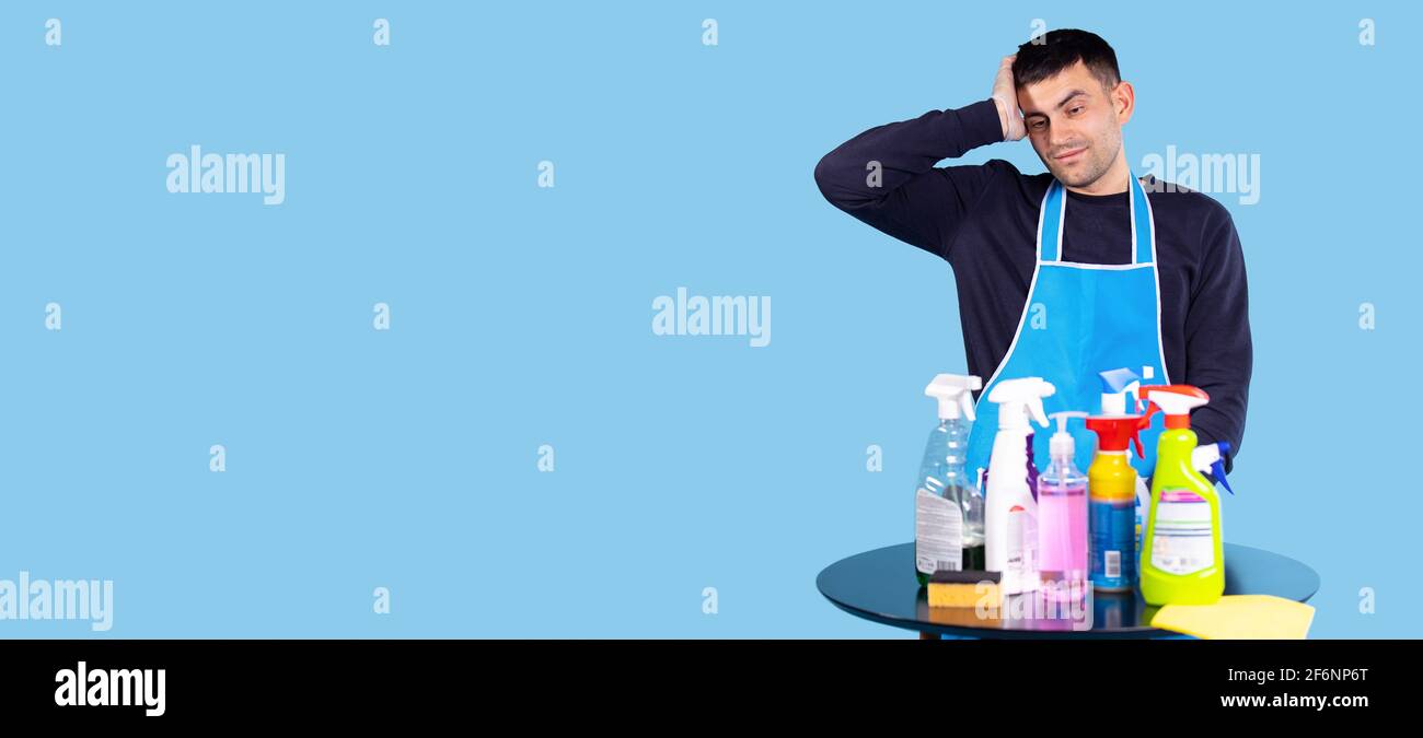 Banner, formato lungo. Giovane uomo stanco di faccende domestiche. Un uomo tiene la mano sulla testa davanti a lui un tavolo di legno con diverse pulizie Foto Stock