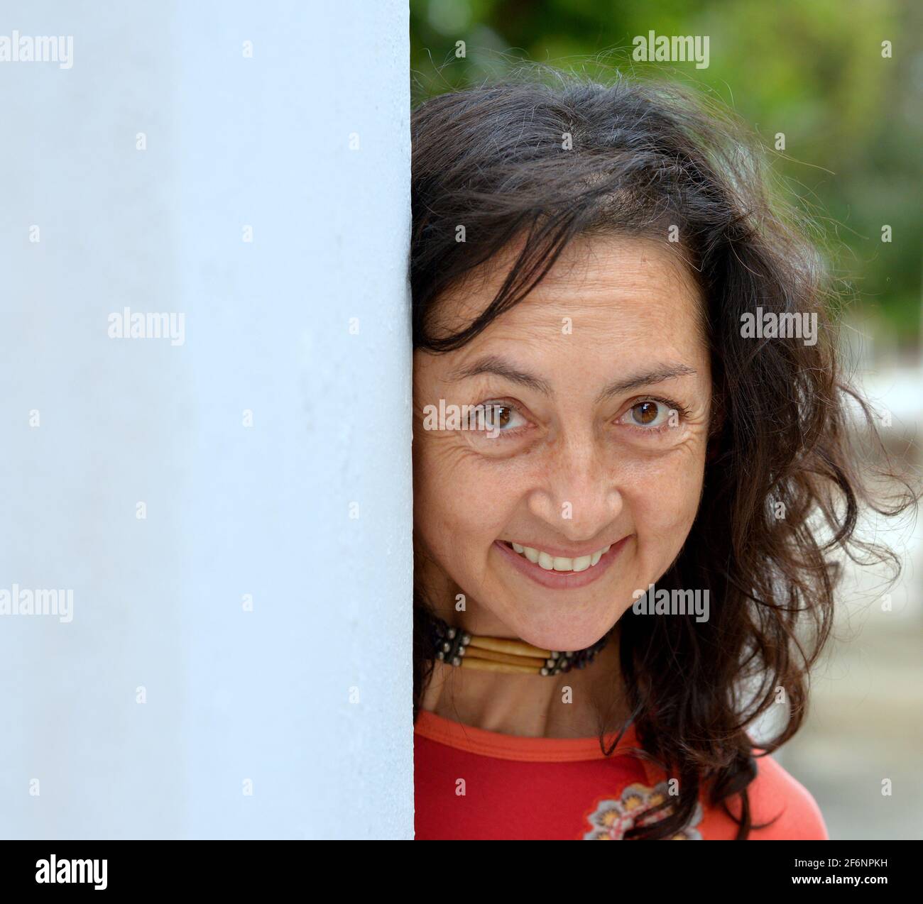 Affascinante donna bruna caucasica matura con lunghi capelli sbirciati intorno a una parete grigia e rende una felice espressione facciale di fronte a uno sfondo parco. Foto Stock