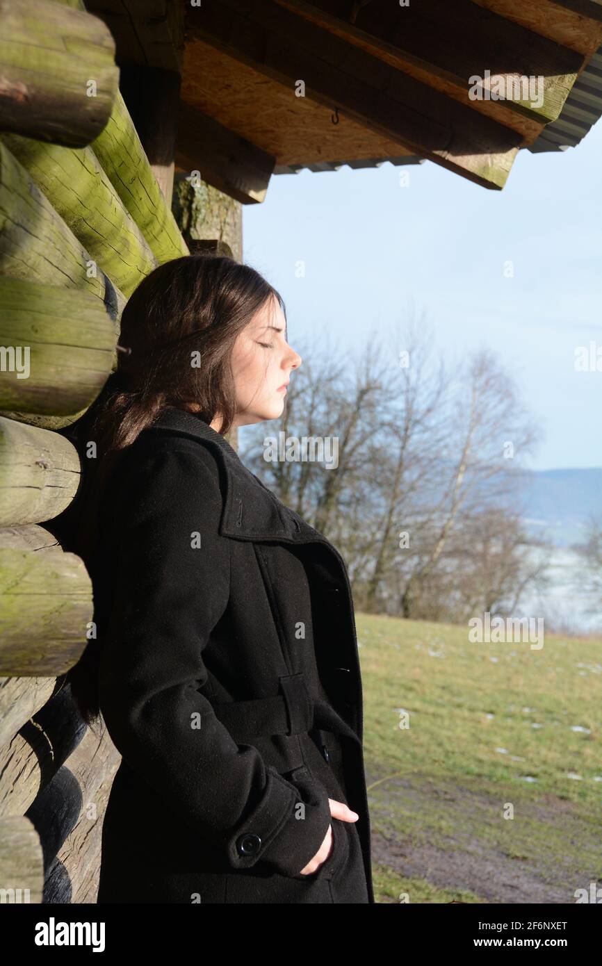 Giovane donna dal lato, con giacca nera, appoggiata contro una capanna di legno, con una seria espressione sul viso e gli occhi chiusi, godendo il sole, io Foto Stock