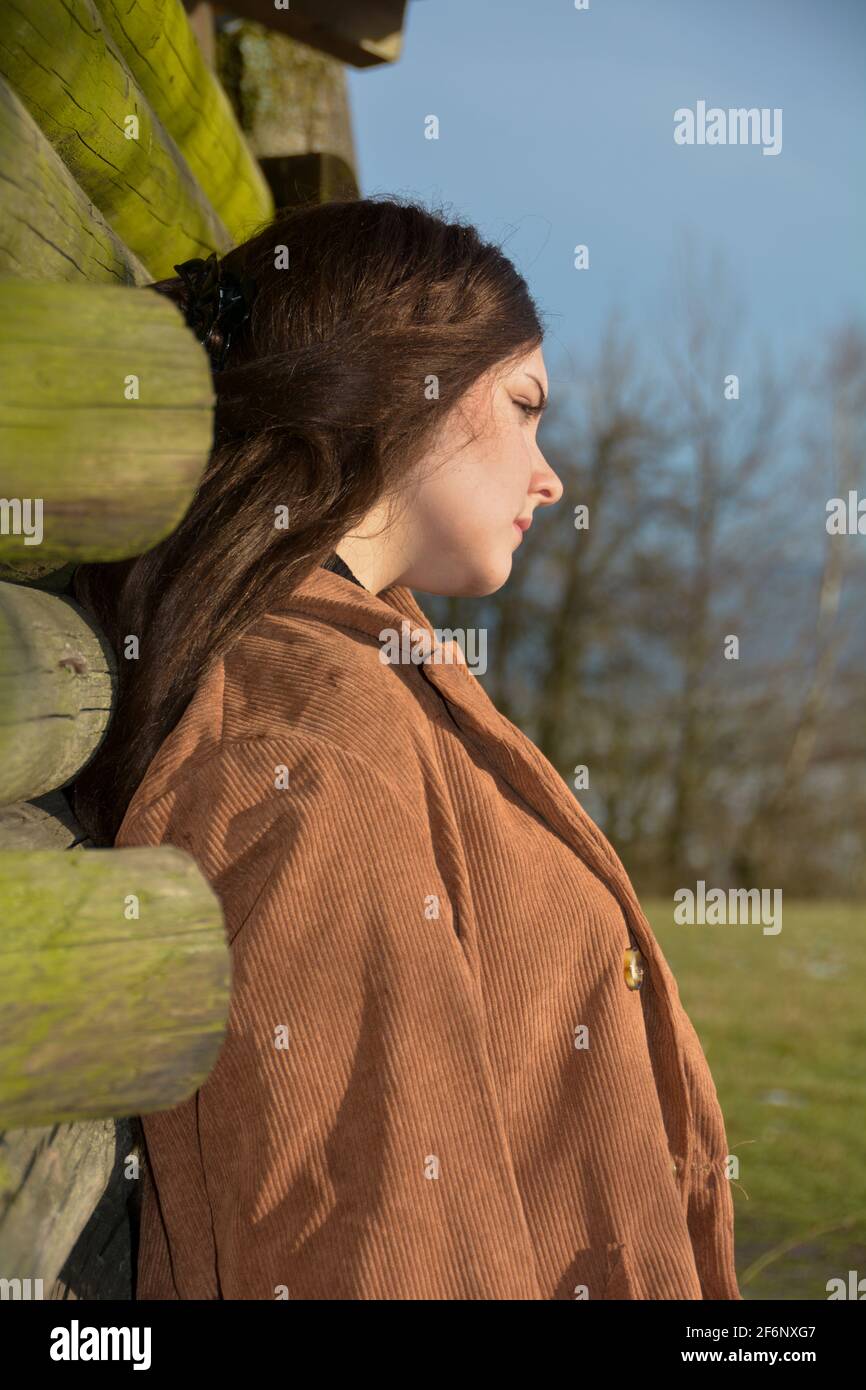 Giovane donna di lato, appoggiata ad una capanna di legno, con un'espressione seria sul viso e gli occhi chiusi, godendosi il sole, in natura verde Foto Stock