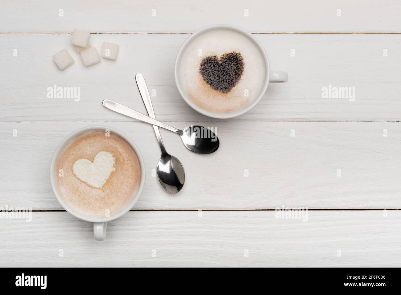 vista dall'alto delle tazze con cappuccino e latte vicino ai cucchiai e cubetti di zucchero su sfondo di legno bianco Foto Stock