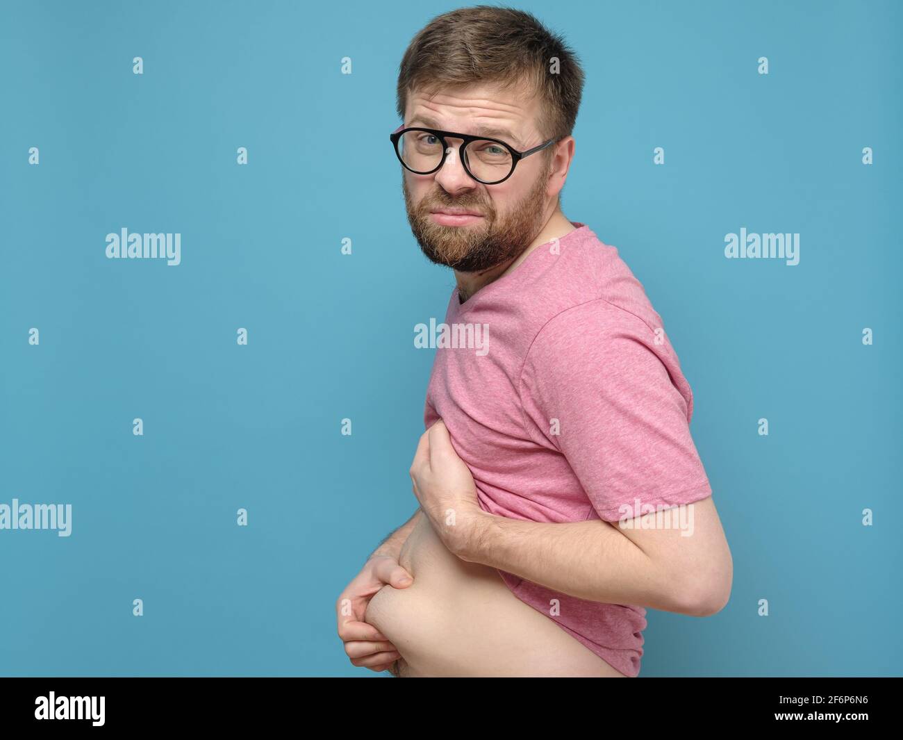 L'uomo sconvolto tiene con le mani il grasso in eccesso sullo stomaco. Persona in sovrappeso. Concetto di nutrizione adeguata e di uno stile di vita sano. Foto Stock