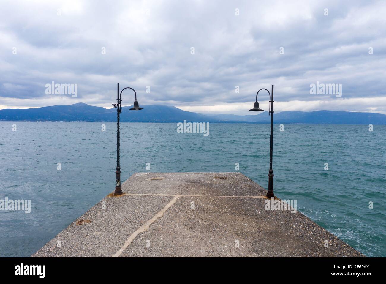 Pier vuoto con luci di strada arrugginite conduce al mare. Cielo nuvoloso e montagne sullo sfondo. Oropos mare sfondo. Golfo di Evia, Grecia. Foto Stock