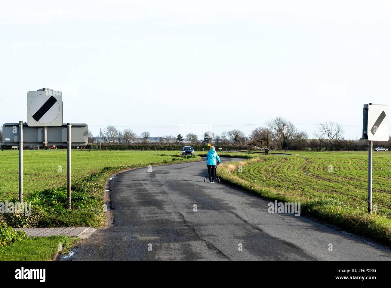 Scena comica, donna con un camminatore elettrico, a piedi lungo una stretta strada di campagna con 2 cartelli "limite di velocità nazionale". Auto che viene verso di lei. Foto Stock