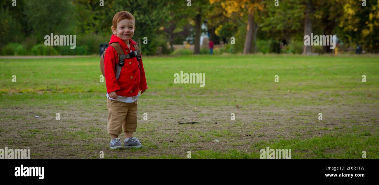 Un bambino carino, rosso, che indossa una giacca rossa in piedi su un prato in un parco in una serata soleggiata Foto Stock