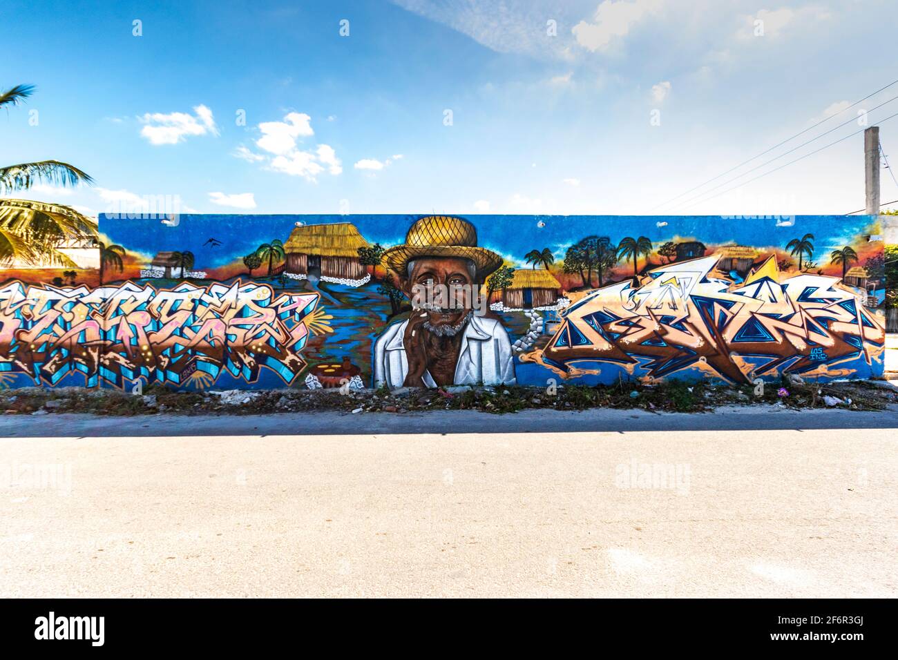 Arte di strada in una piccola città su una lunga parete con un vecchio che indossa un cappello di paglia, una casetta rustica e parole, con un cielo blu sopra la parete Foto Stock