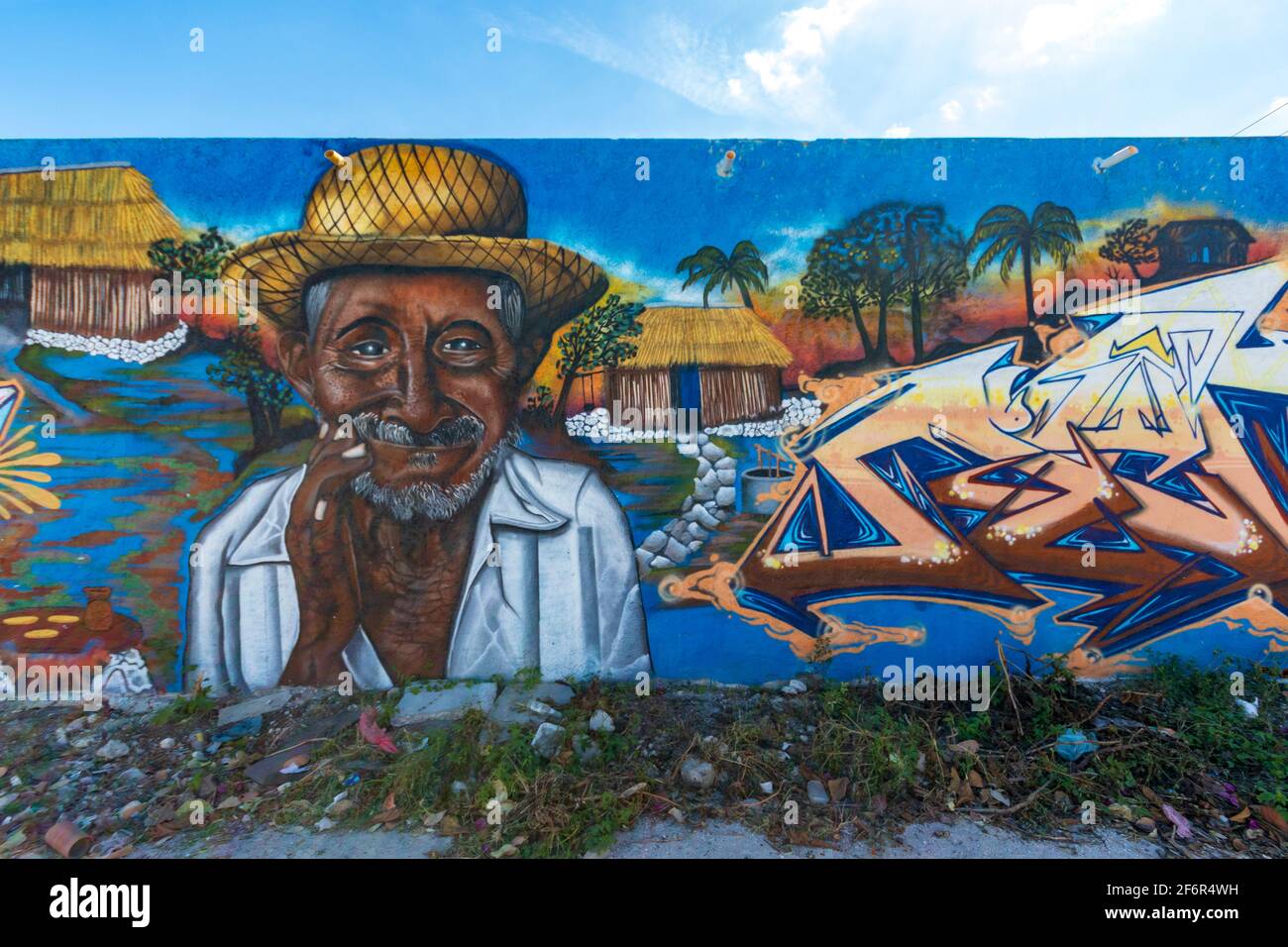 Arte di strada in una piccola città su una lunga parete con un vecchio che indossa un cappello di paglia, una casetta rustica e parole, con un cielo blu sopra la parete Foto Stock