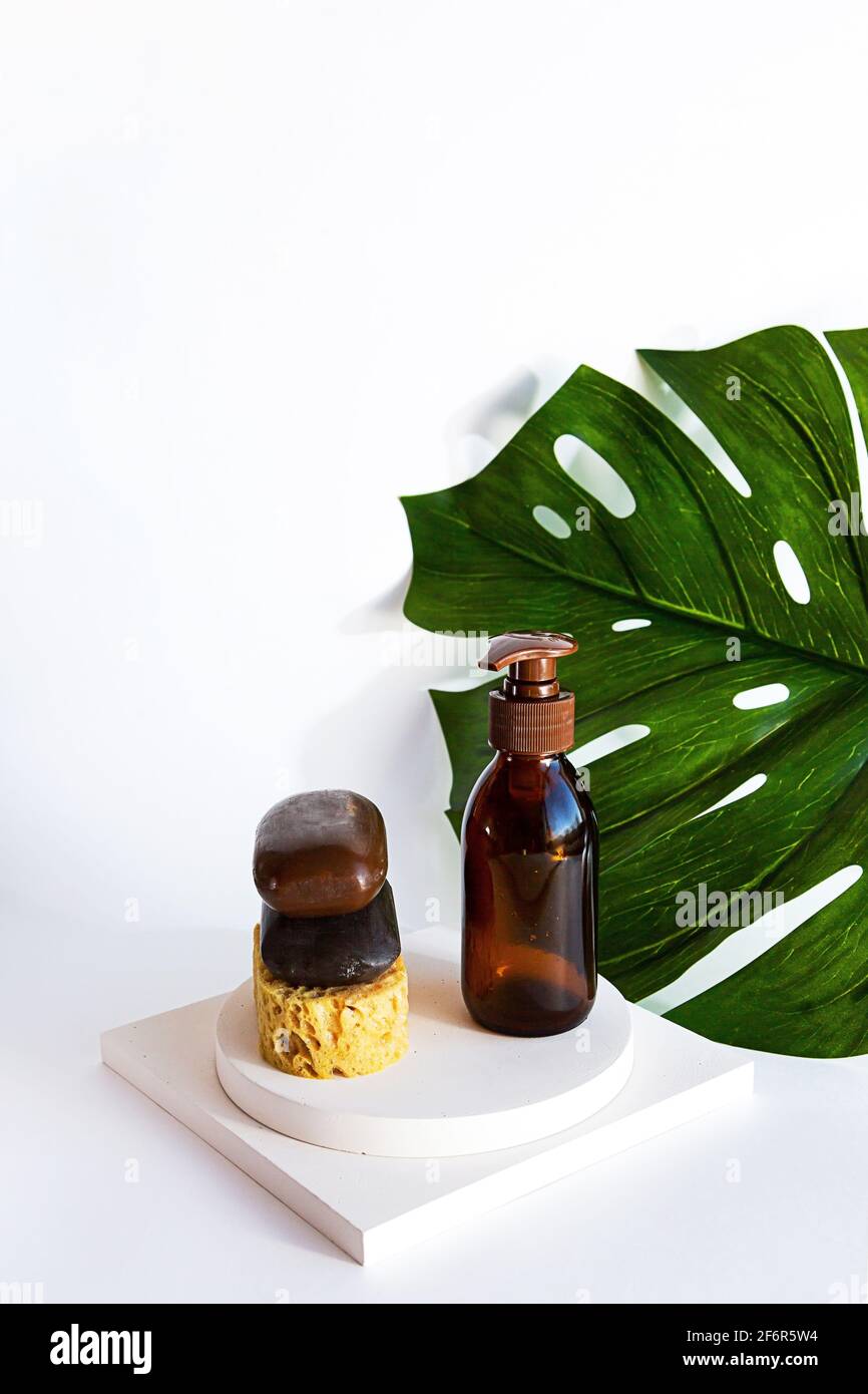 Saponette naturali fatte a mano e shampoo biologico in una bottiglia di vetro. Prodotti per la cura del corpo e delle foglie di palma Monstera su sfondo bianco. Foto Stock