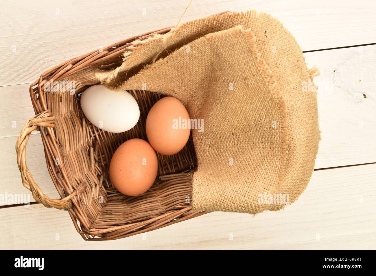 Tre uova di pollo fresche, biologiche, deliziose, beige e bianche in un cesto di vimini con un tovagliolo di iuta su un ripiano in legno bianco. Foto Stock