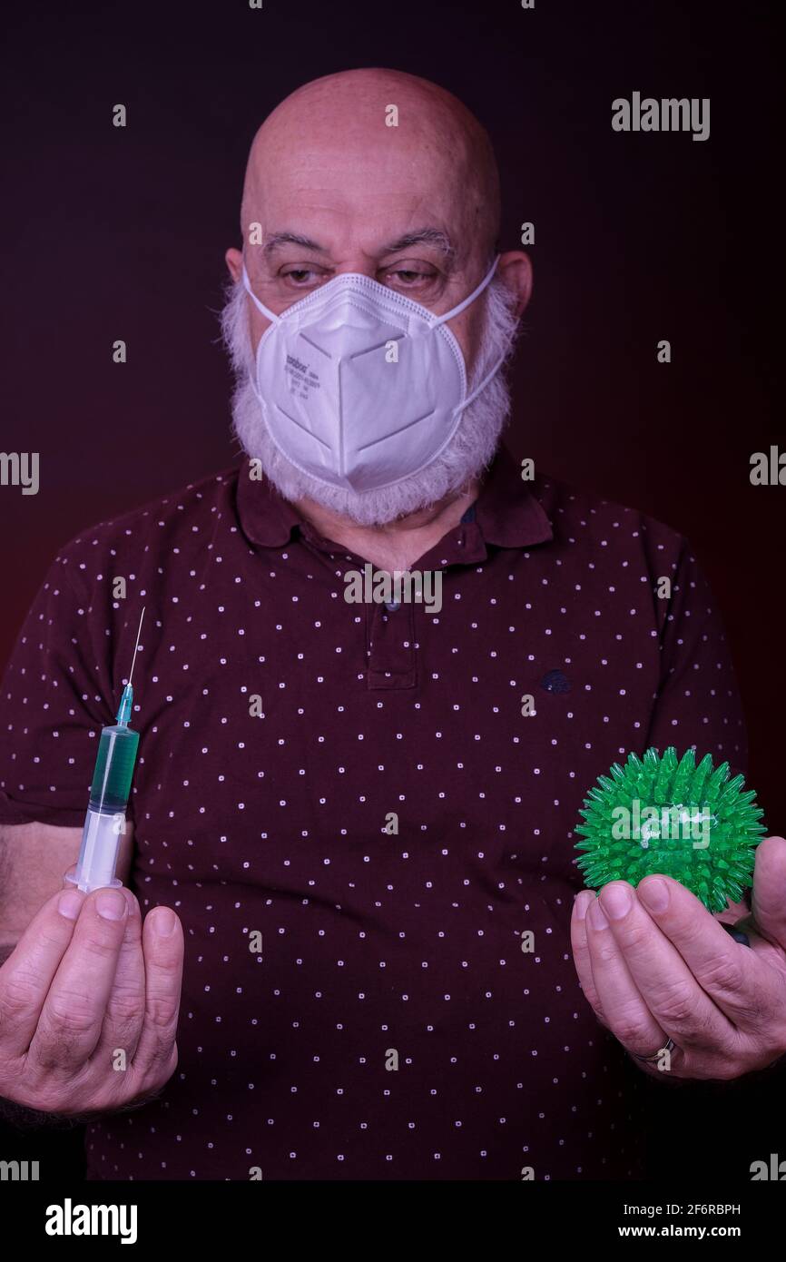 l'uomo anziano con maschera protettiva contiene una siringa di vaccino una mano e una palla verde che rappresenta il virus corona in un altro Foto Stock