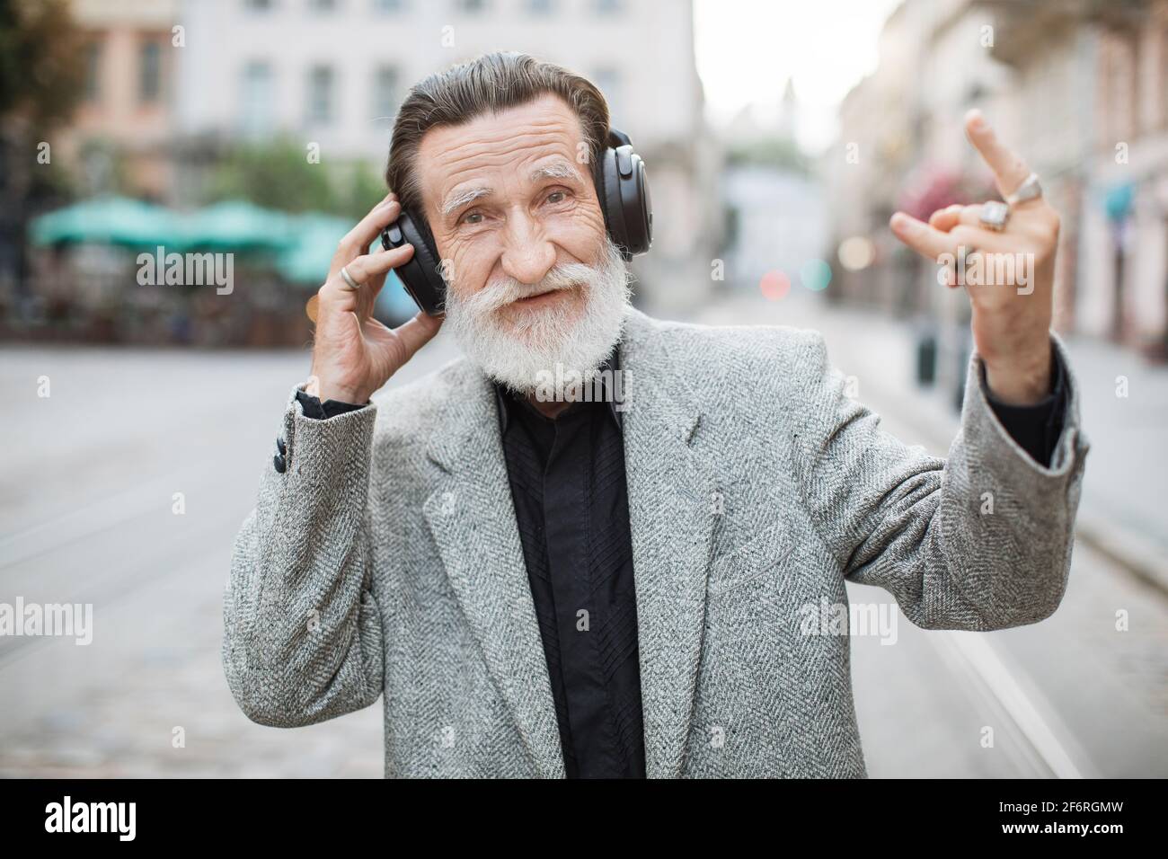 Uomo anziano divertente con la barba grigia che indossa le cuffie wireless  e mostra il simbolo della roccia mentre si trova in città. Buon pensionato  che ascolta musica all'aperto Foto stock -