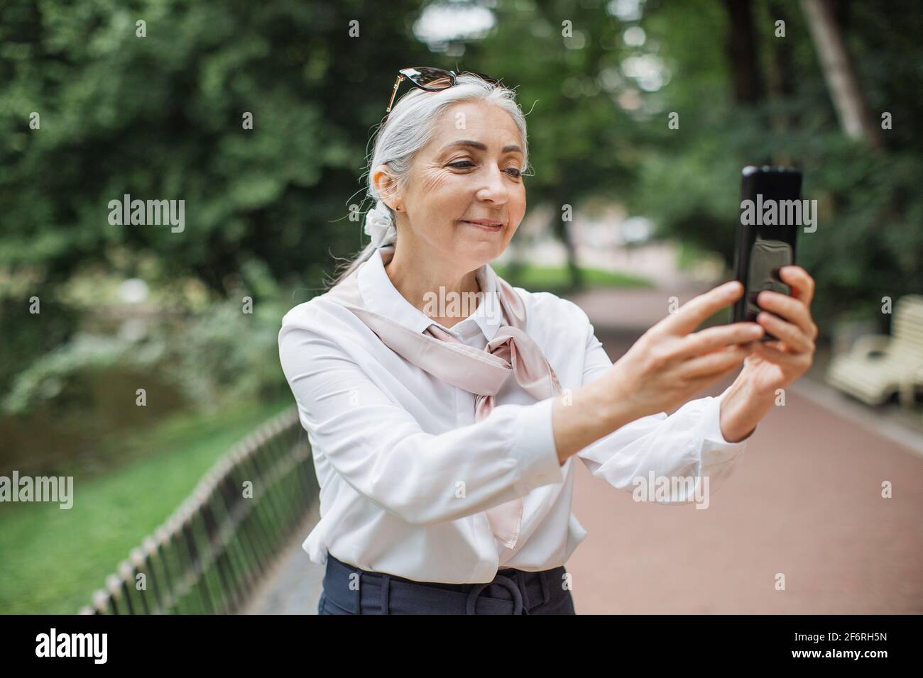 Donna anziana affascinante in abiti eleganti con smartphone mentre si  cammina nel parco verde. Signora matura con occhiali da sole sulla testa in  piedi all'aperto con gadget moderni in mani Foto stock -