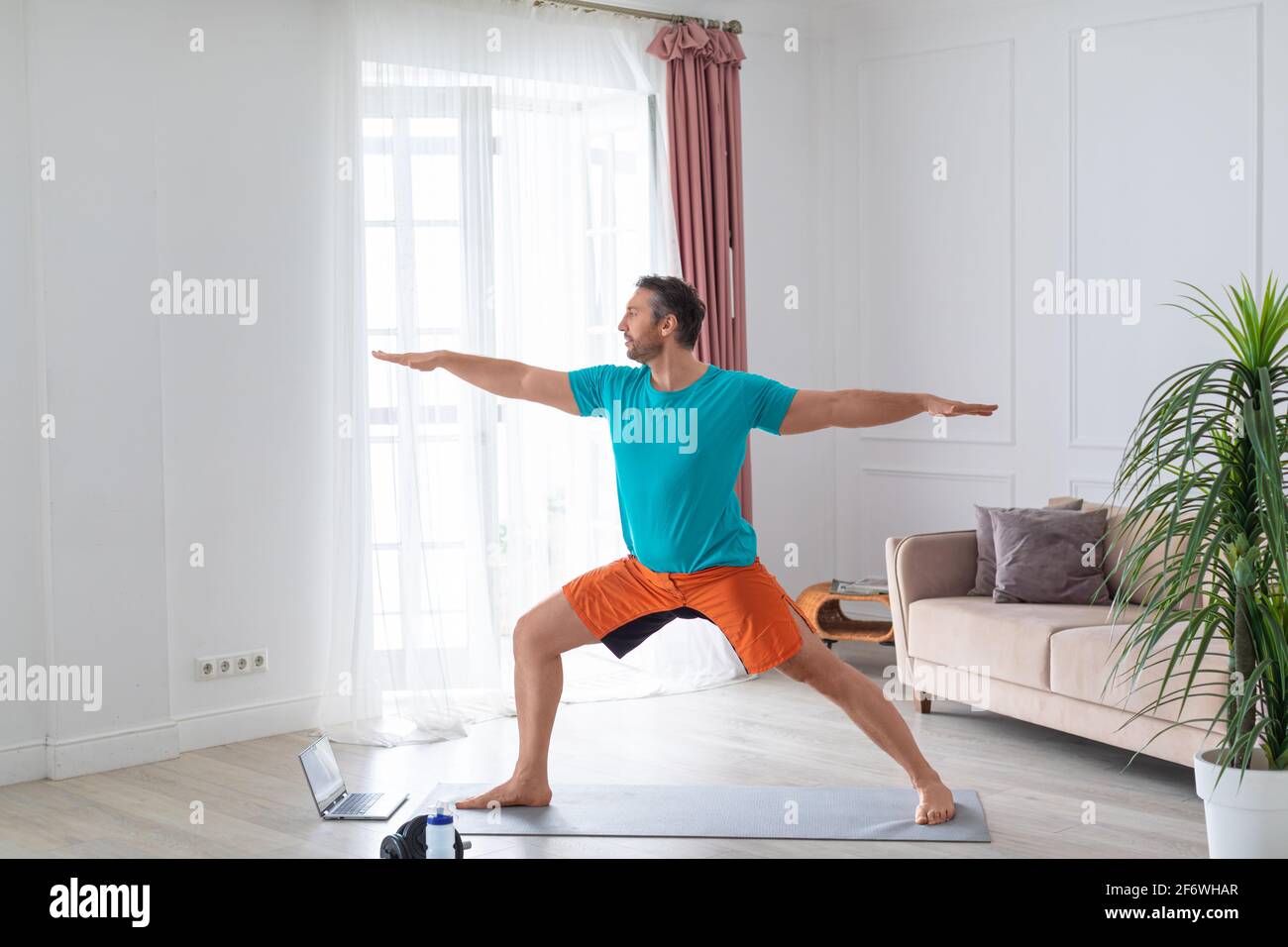 L'uomo medio anziano lavora a casa con un tutorial online. Esegue un esercizio di equilibrio davanti a un monitor laptop in casa. Foto Stock