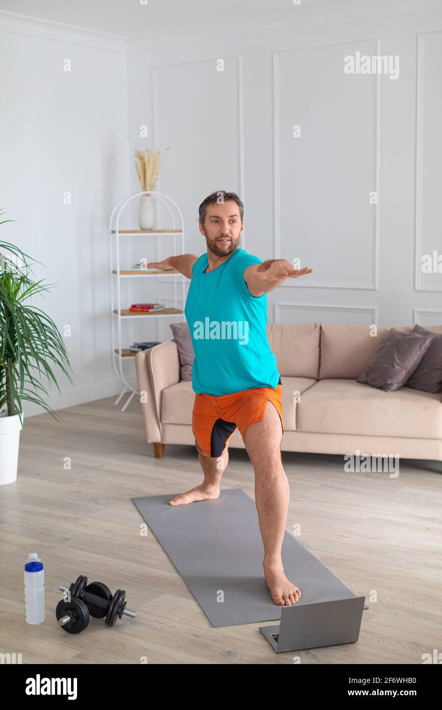 Un uomo di mezza età lavora a casa con un tutorial online. Esegue un esercizio di equilibrio davanti a un monitor portatile. Foto verticale. Foto Stock