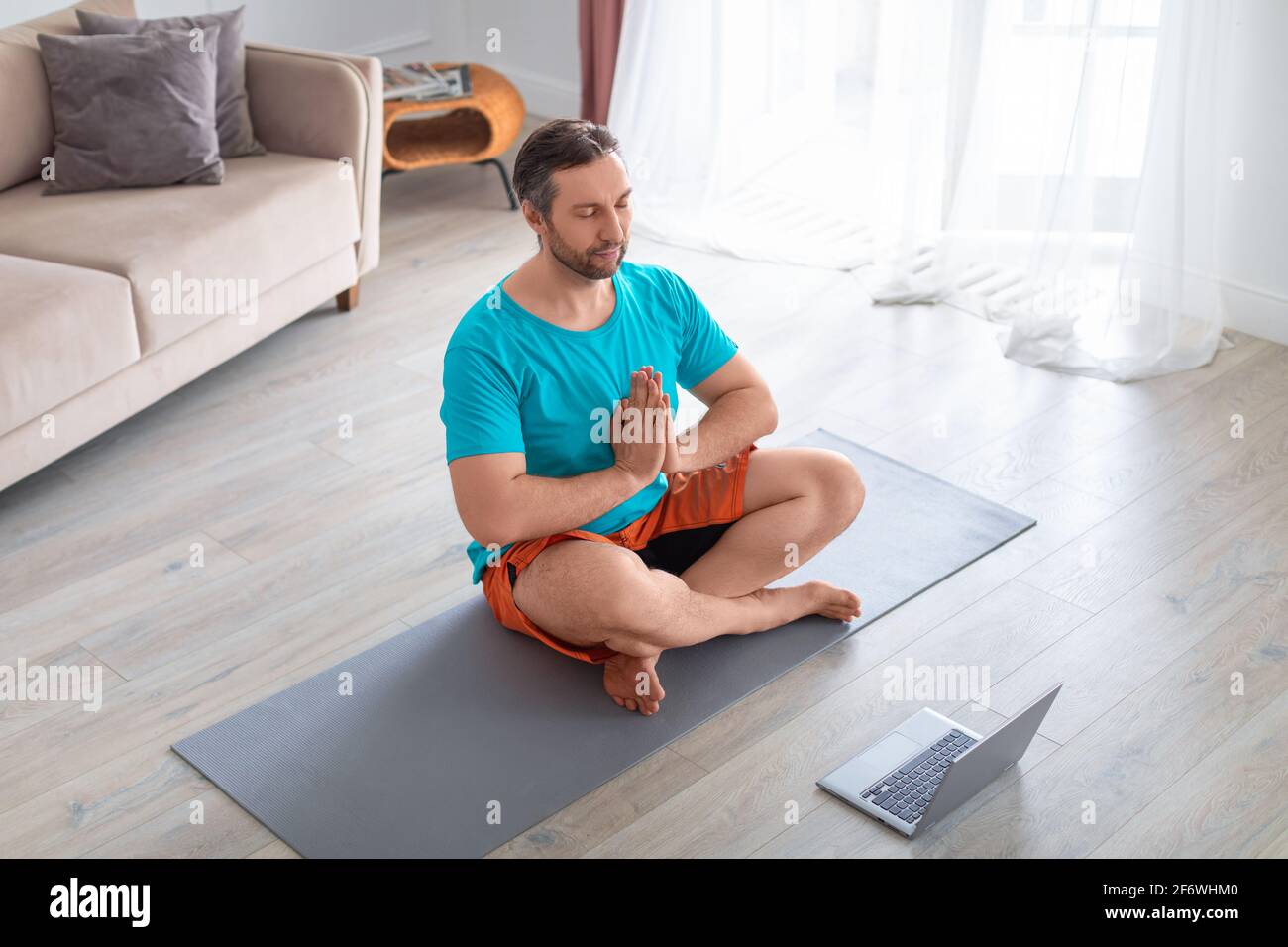 Uomo medio anziano meditando davanti al monitor del computer portatile. Allenamento di yoga domestico. Foto Stock