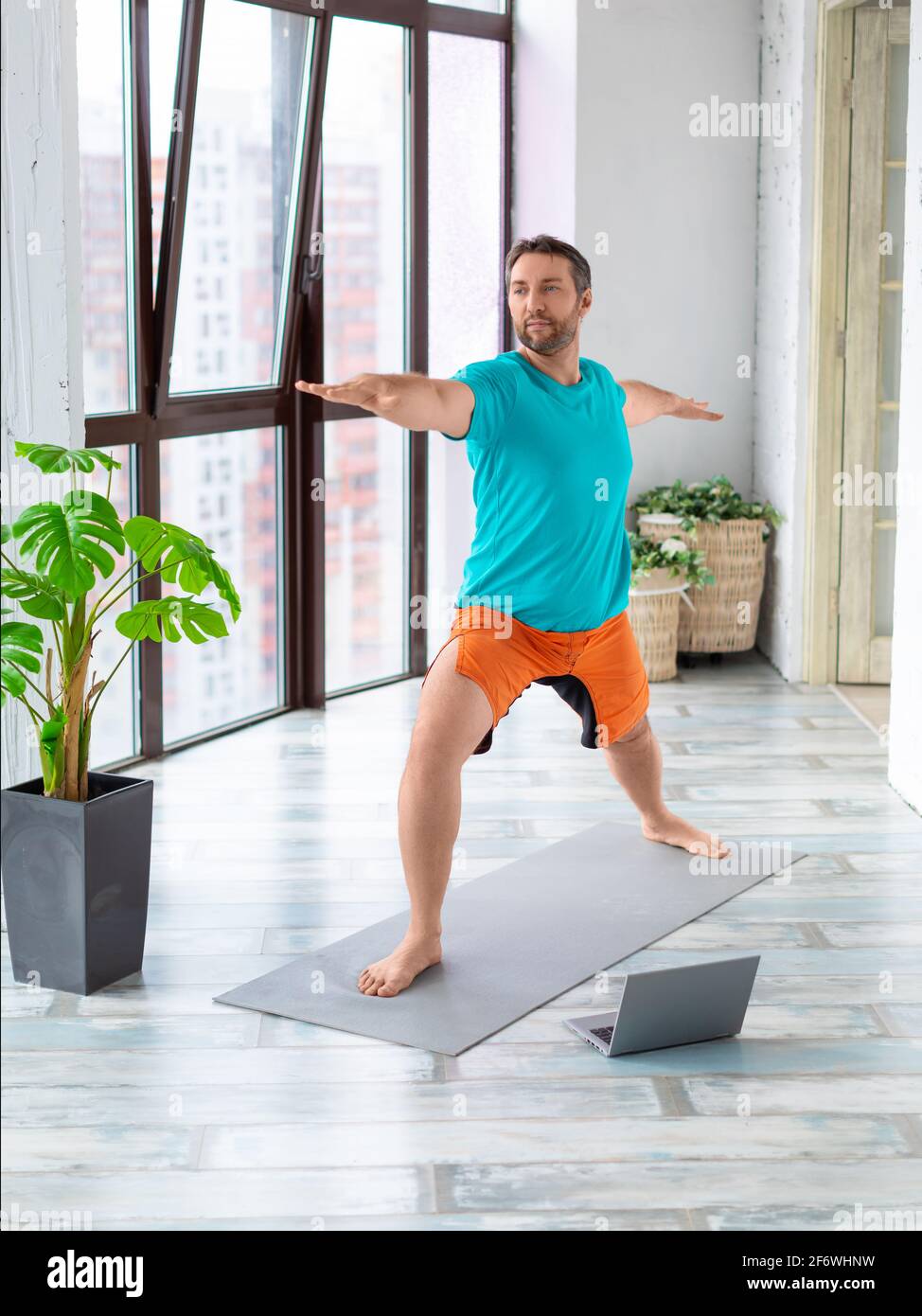Un uomo di mezza età si sta esercitando a casa. Esegue un esercizio di equilibrio davanti a un monitor portatile. Corsi di formazione online con un tutorial dall'interno Foto Stock