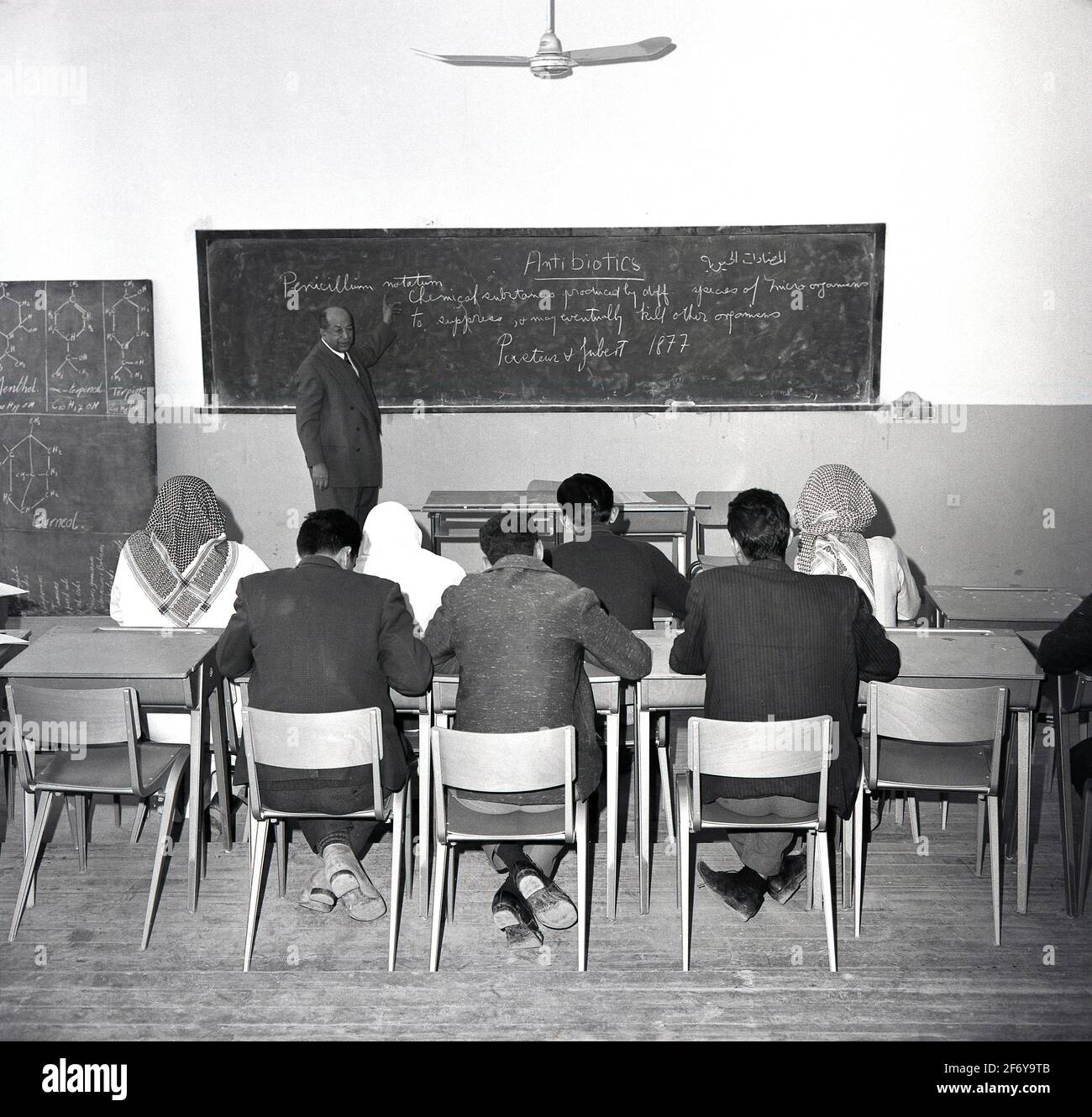 Anni '60, storici, studenti arabi di sesso maschile, alcuni indossano sciarpe alla testa, seduti in una classe con un insegnante di sesso maschile alla lavagna, Jeddah, Arabia Saudita. È interessante notare che la lezione sembra essere in inglese, poiché la scrittura sulla lavagna relativa agli antibiotici è in lingua inglese. Foto Stock