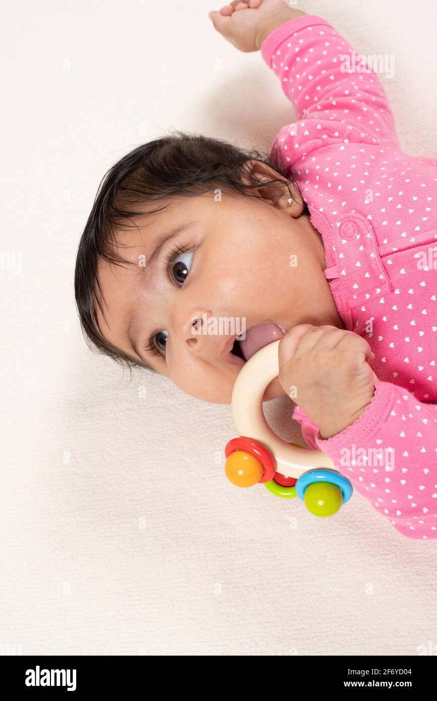 4 mesi bambina vestire ragazza sul retro guardando a lato come si bocca giocattolo di legno, lingua visibile Foto Stock