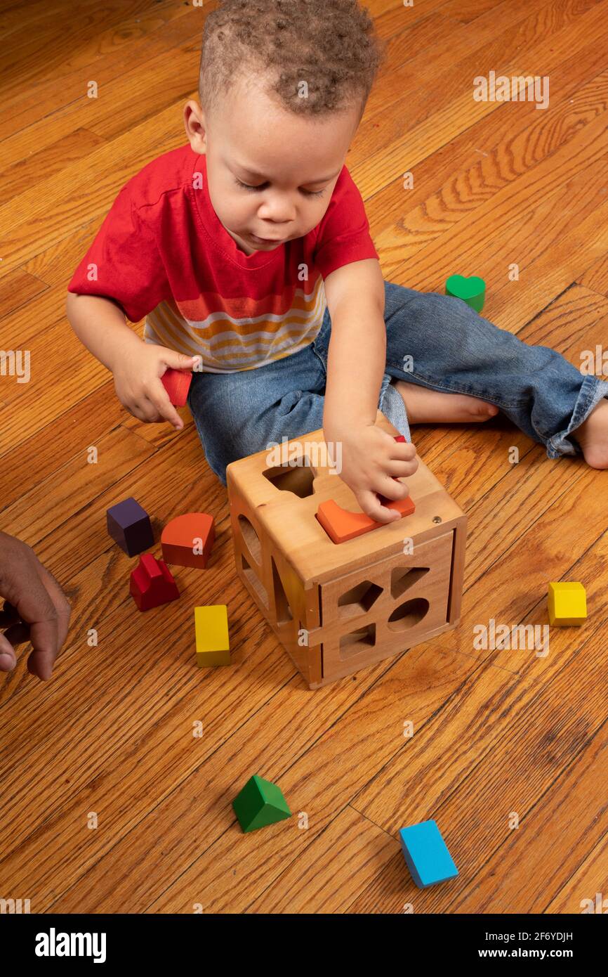 Ragazzo di due anni che gioca con il giocattolo dello smistatore di forma in legno, girando pezzo per inserirsi nella fessura Foto Stock