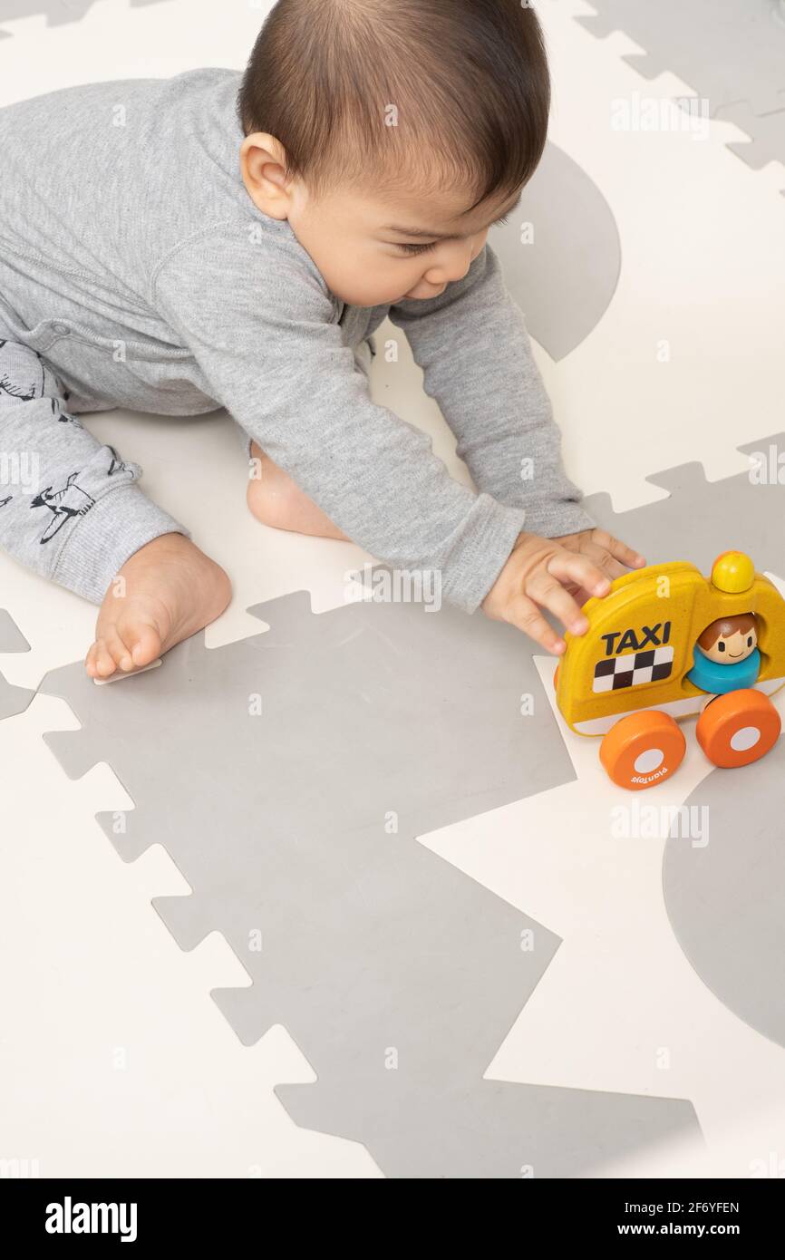 Bambino di sei mesi che si siede appoggiato sopra per raggiungere il giocattolo, un veicolo di taxi di legno Foto Stock