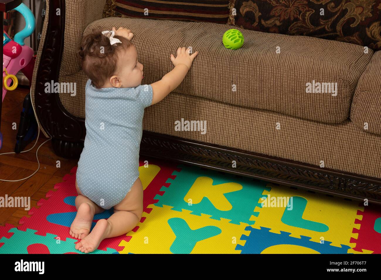 bambina di 7 mesi, inginocchiata sul divano interessata alla palla sul cuscino, pronta a tirarsi in piedi per raggiungerla Foto Stock